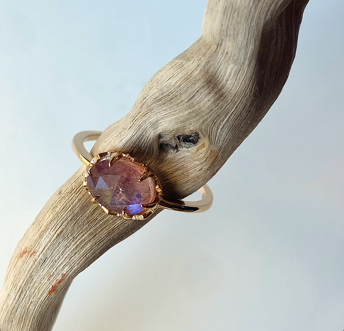 Lula Sea Jewellery New Zealand, Sugarplum ring, ring is pictured on a pice of drift wood, showing the blue flash of the strawberry labradorite faucet cut gemstone. Ring made from Gold vermeil