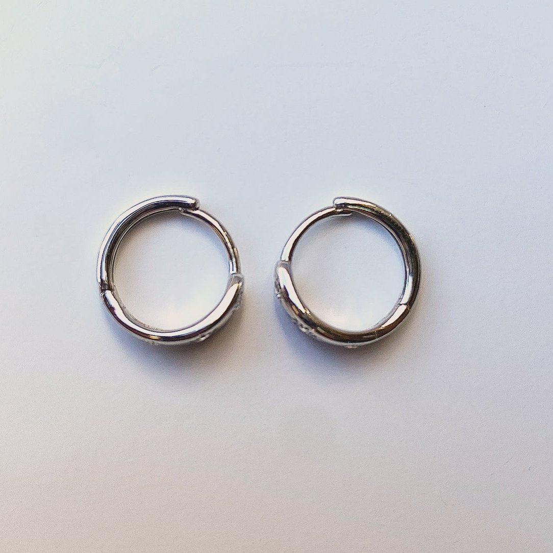 Lula Sea Jewellery Nz, Sun Hoop Earrings in sterling silver, huggie hoops featuring three radiant cubic zirconia suns, each with finely engraved rays that catch the light. Pictured on flat surface from above 